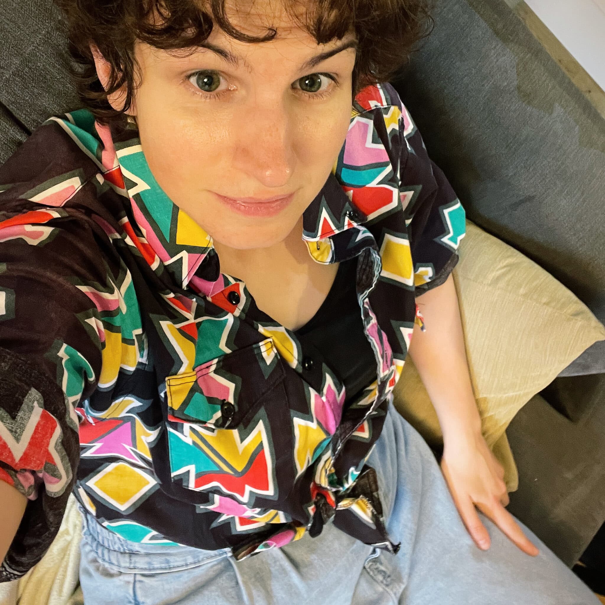 Alex, a white non-binary person in their 30s wearing a colourful shirt looks up at the camera with a bit of curly hair on their forehead.